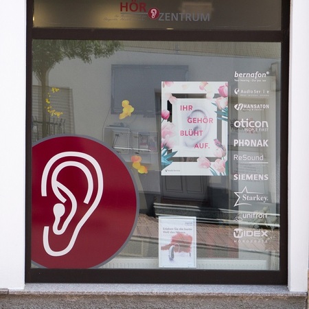 Schaufenster eines Hörakustik-Geschäfts mit großem Ohr-Symbol und Hörgerätehersteller-Logos.